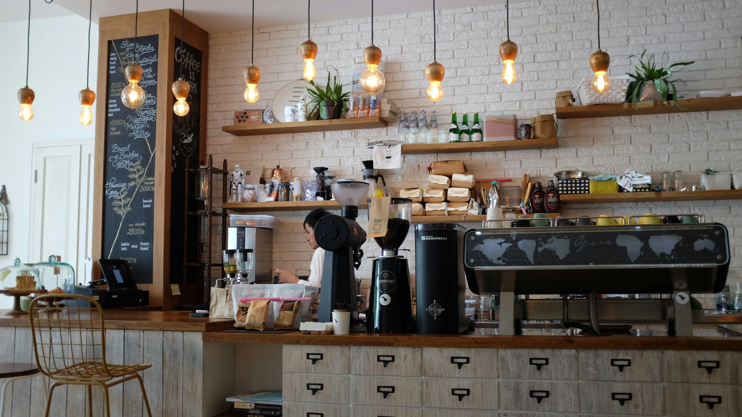 Coffee shop with white brick walls, pendant lighting, and barista working behind wooden counter with espresso machine and grinders.