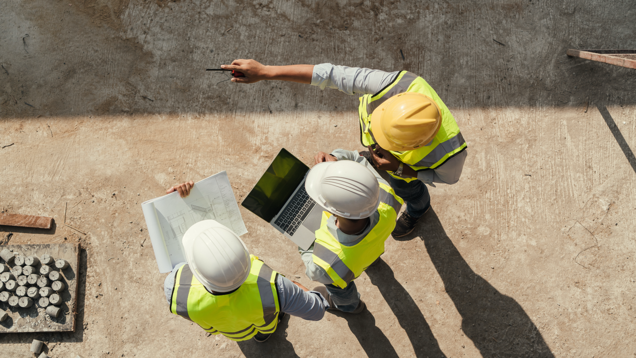 Three construction workers in yellow high-visibility vests and white hard hats standing on a concrete surface, viewed from above. One worker is pointing while the other two look at an open laptop computer and what appears to be architectural plans or blueprints spread on the ground. Construction materials including metal pipes are visible in the background.