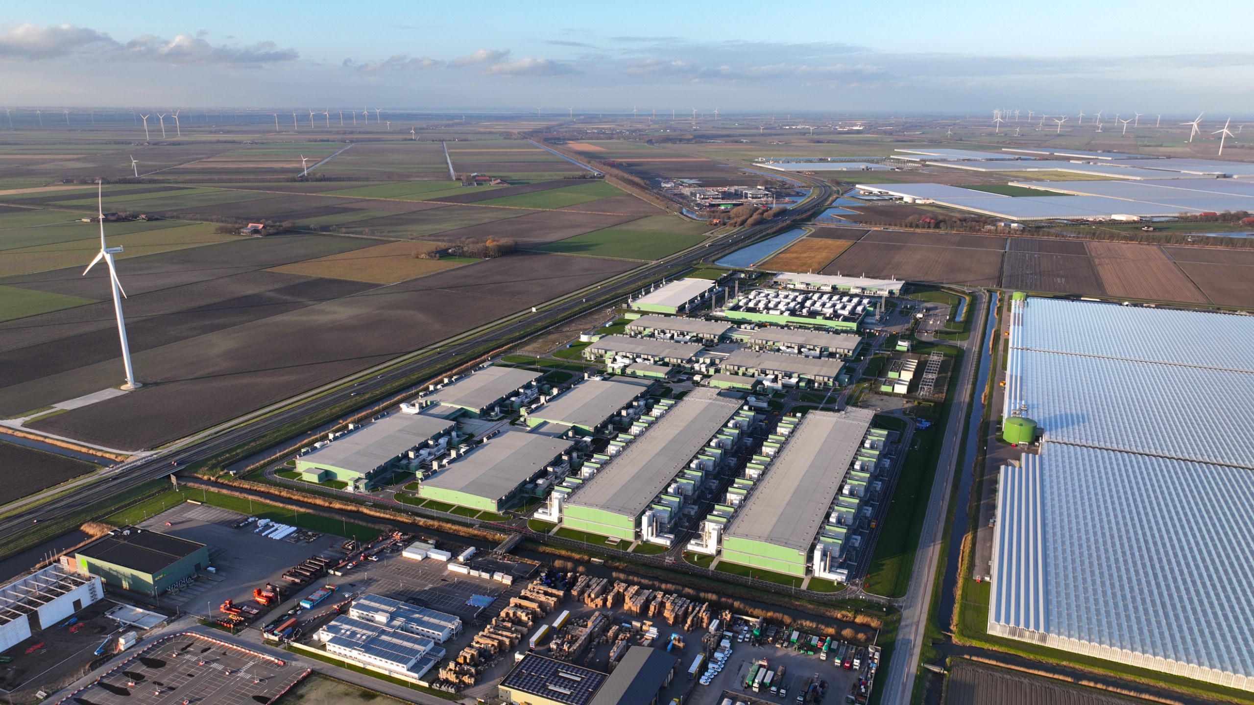 Aerial view of a large data centre complex surrounded by farmland, greenhouses, and wind turbines.