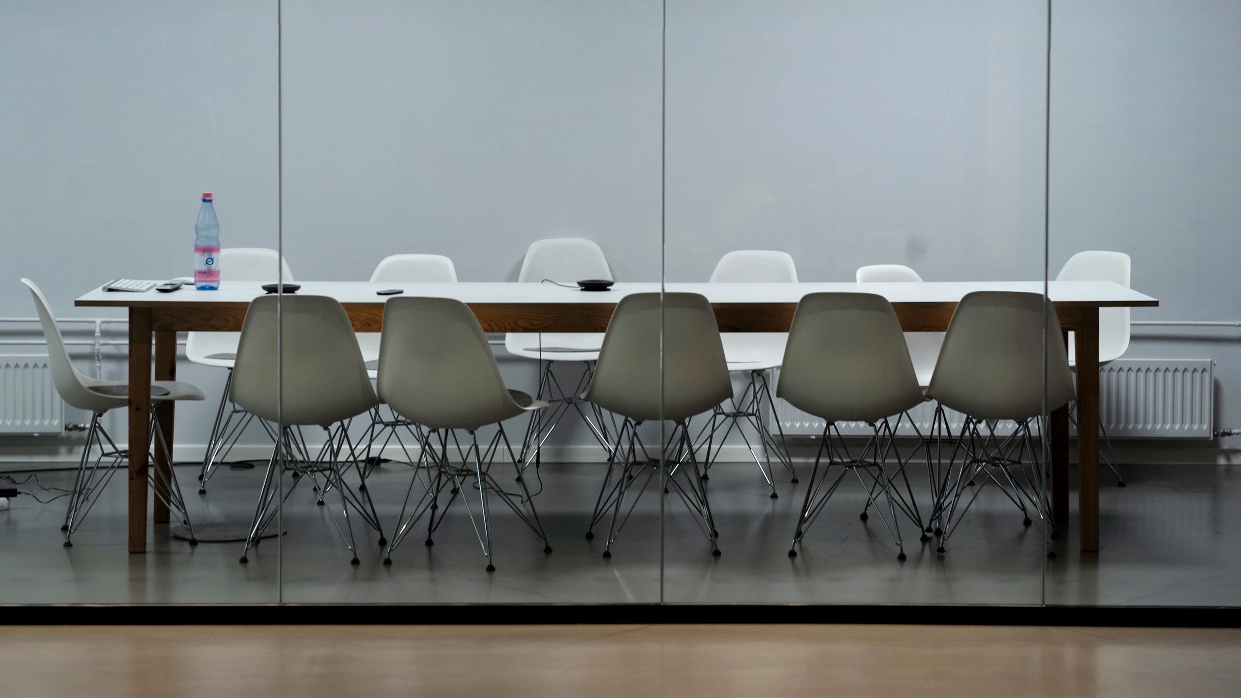 Modern conference room with a long wooden table and a combination of white and taupe Eames-style chairs reflected in a glass partition, a solitary water bottle and notepad on the table indicating a recent or upcoming meeting.