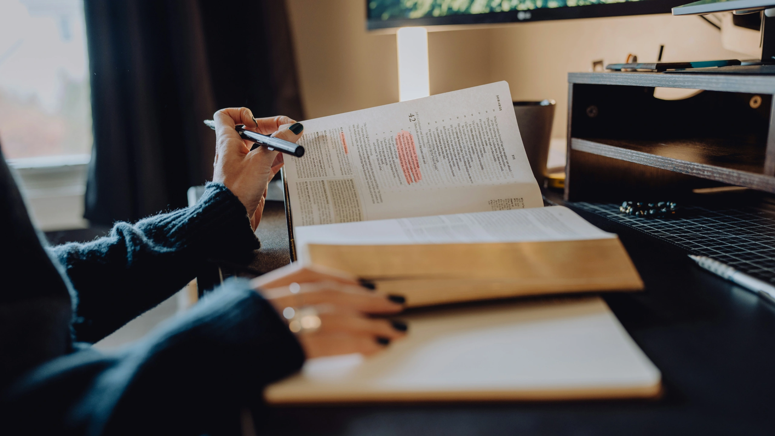 Person sitting at a wooden desk holding an open textbook with highlighted passages in orange and yellow while writing notes. A laptop and coffee cup are visible on the desk.