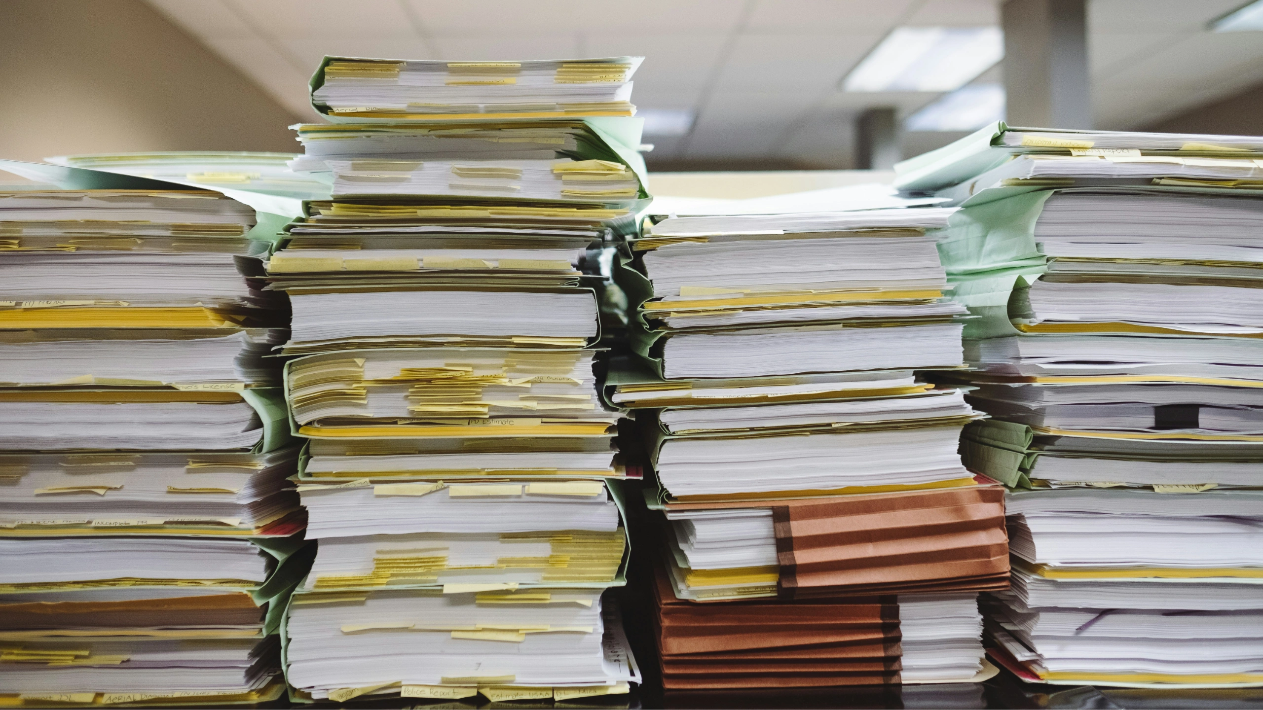 Tall stacks of paper files and folders piled on a desk in an office setting.