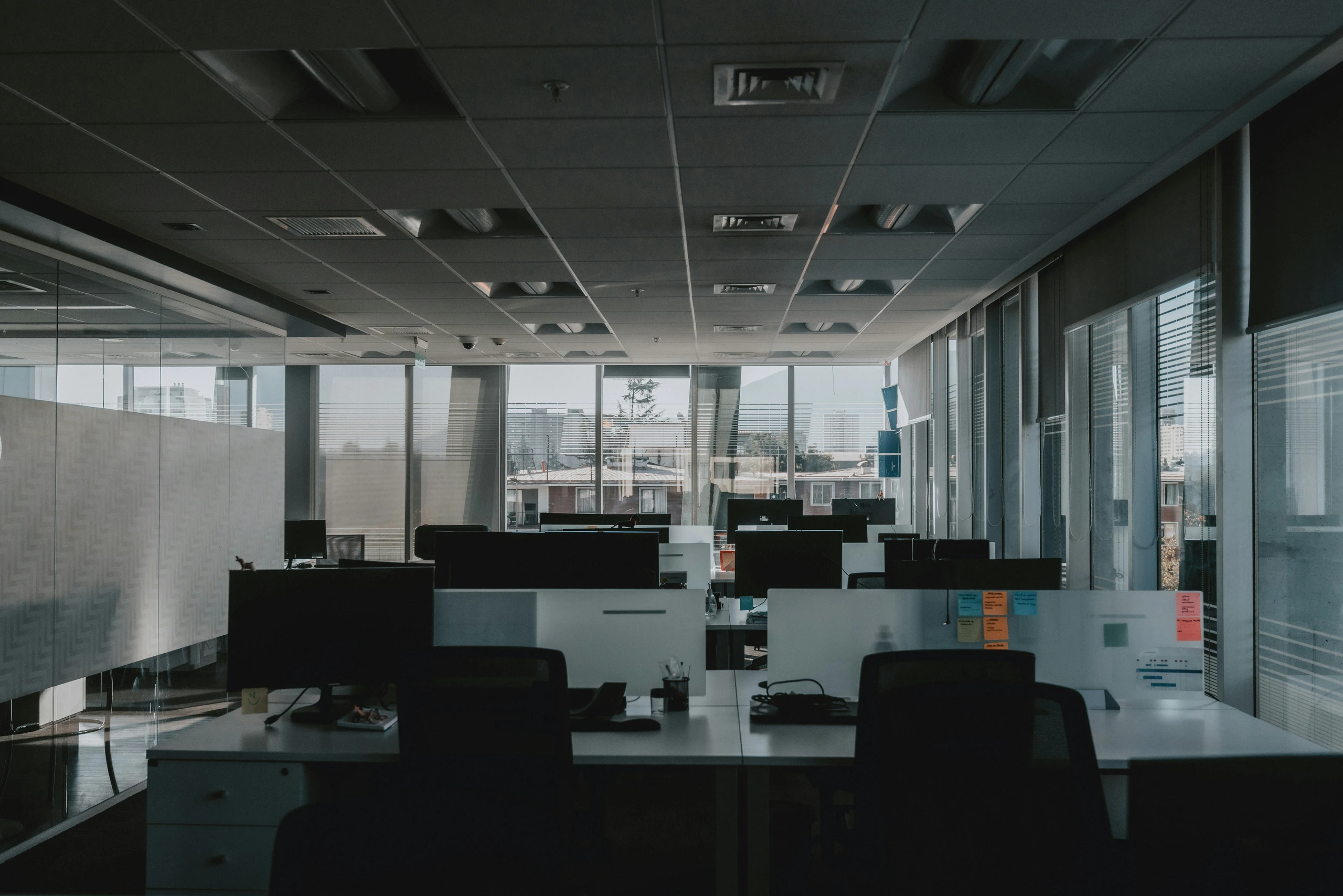 Empty open-plan office with rows of desks, monitors, and chairs, glass partitions, and large windows with horizontal blinds.