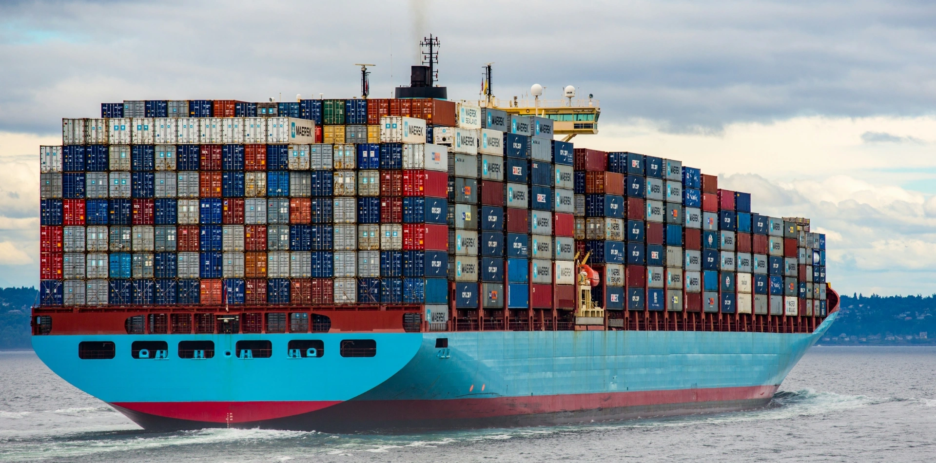 Large Maersk container ship fully loaded with colourful shipping containers, sailing on calm water under overcast sky.