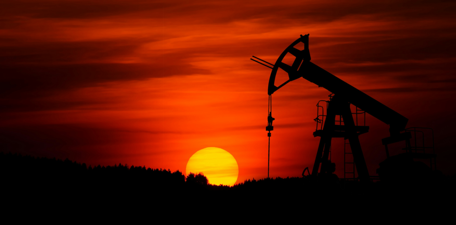 Oil pump jack silhouetted against a red and orange sunset sky, with the sun setting behind a dark treeline.