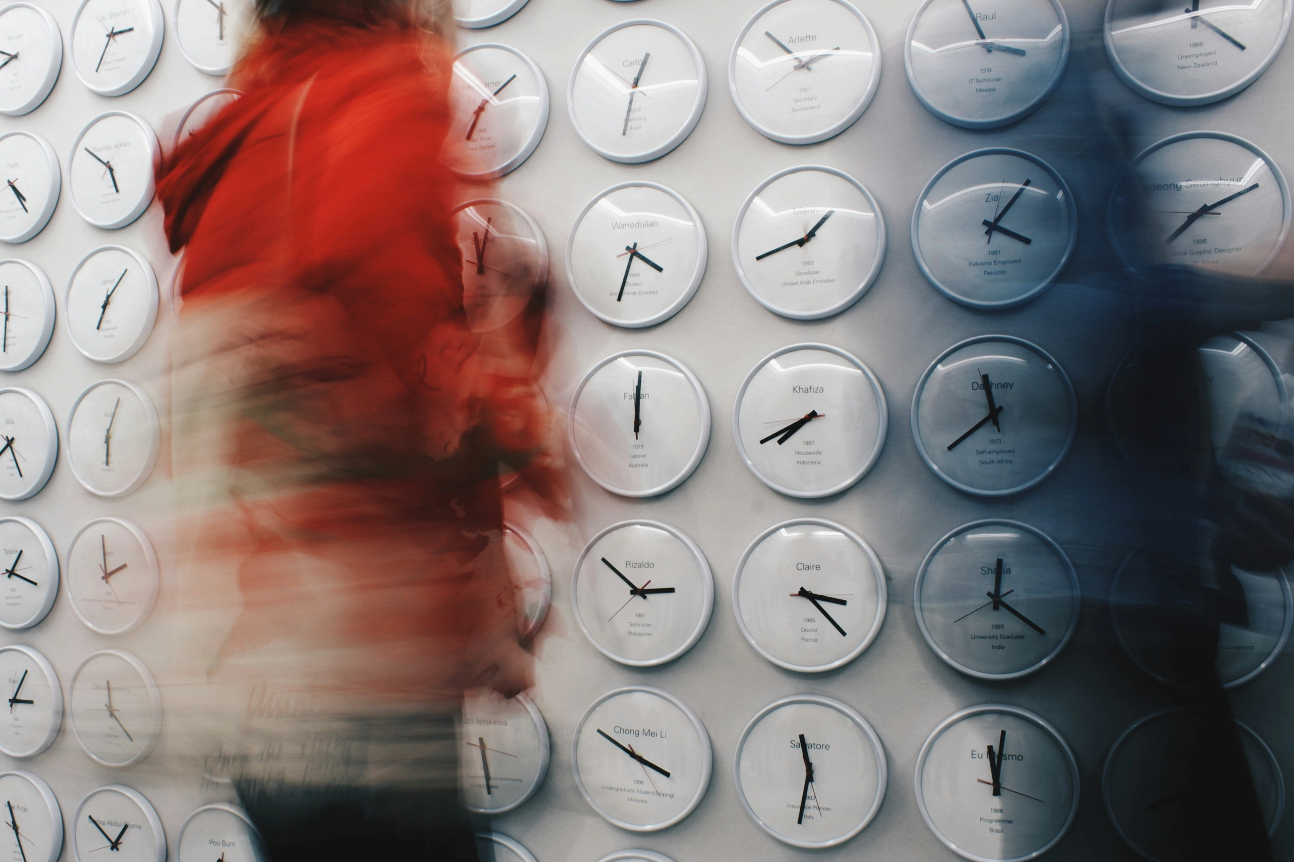 Person in red walking past a wall of white clocks, their figure blurred by motion while the clocks stay sharp.