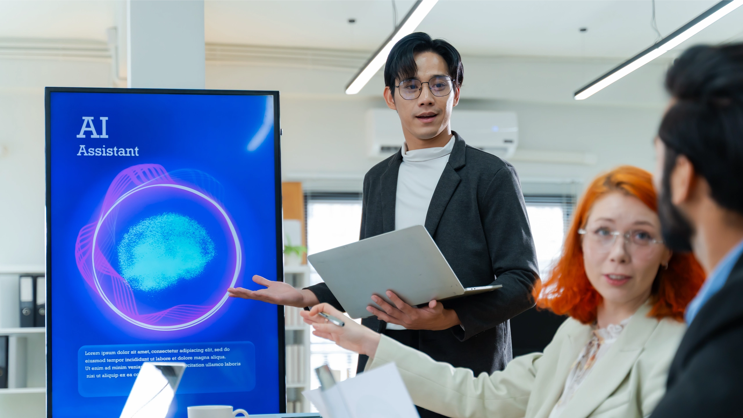 Person in blazer holding laptop presents to group in office; blue digital display showing AI Assistant logo with purple and cyan circular design on desk.