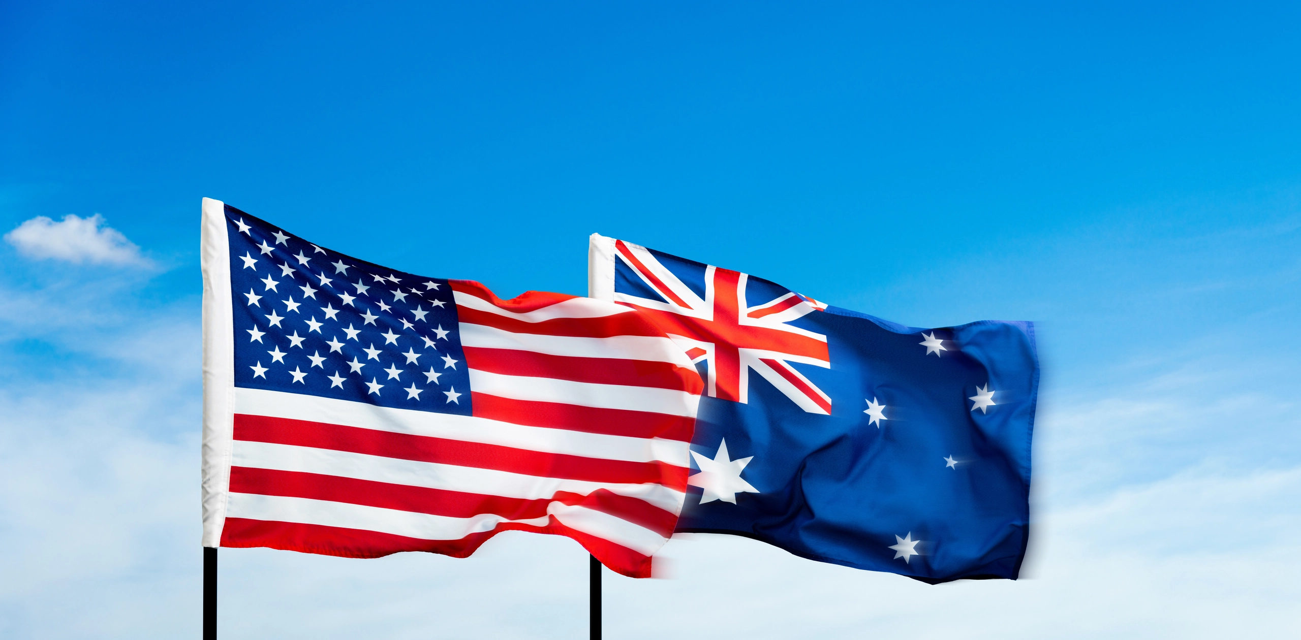 United States flag and Australian flag on flagpoles, flying together against a blue sky with light cloud.