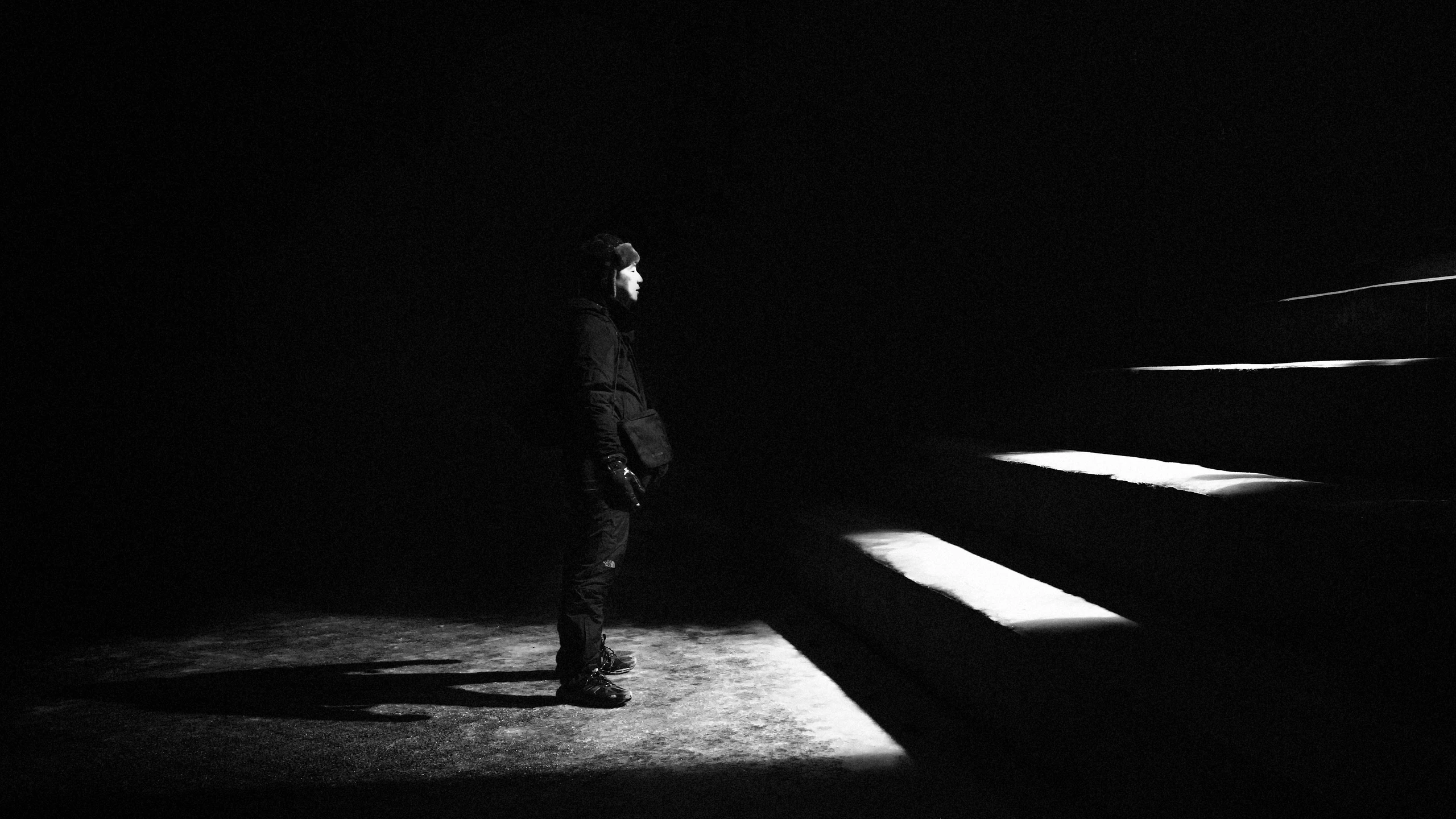 Person in winter clothing stands in darkness, face lit by a beam of light beside illuminated stair edges.