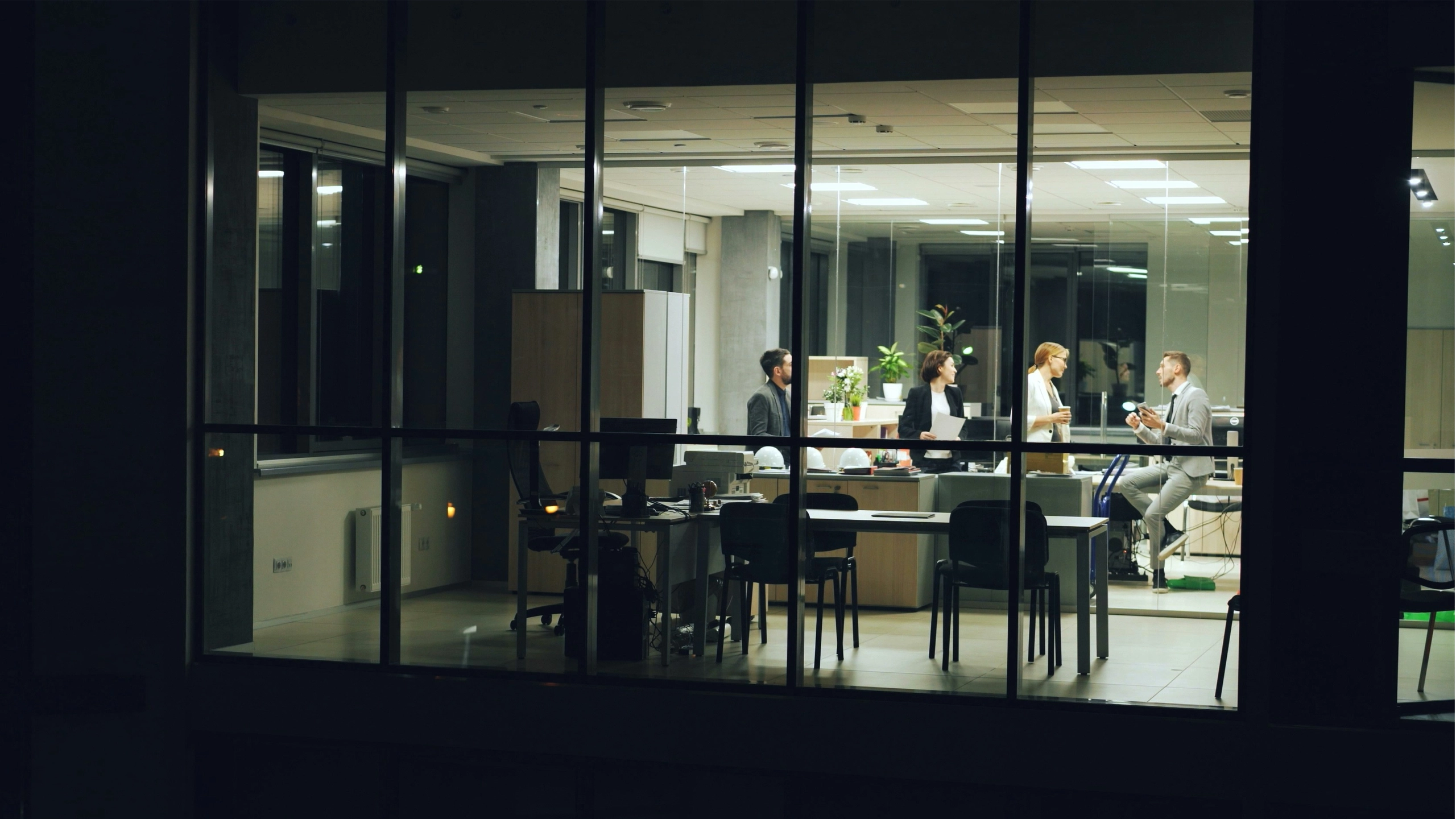 Office at night seen through windows, four people standing near desks talking.