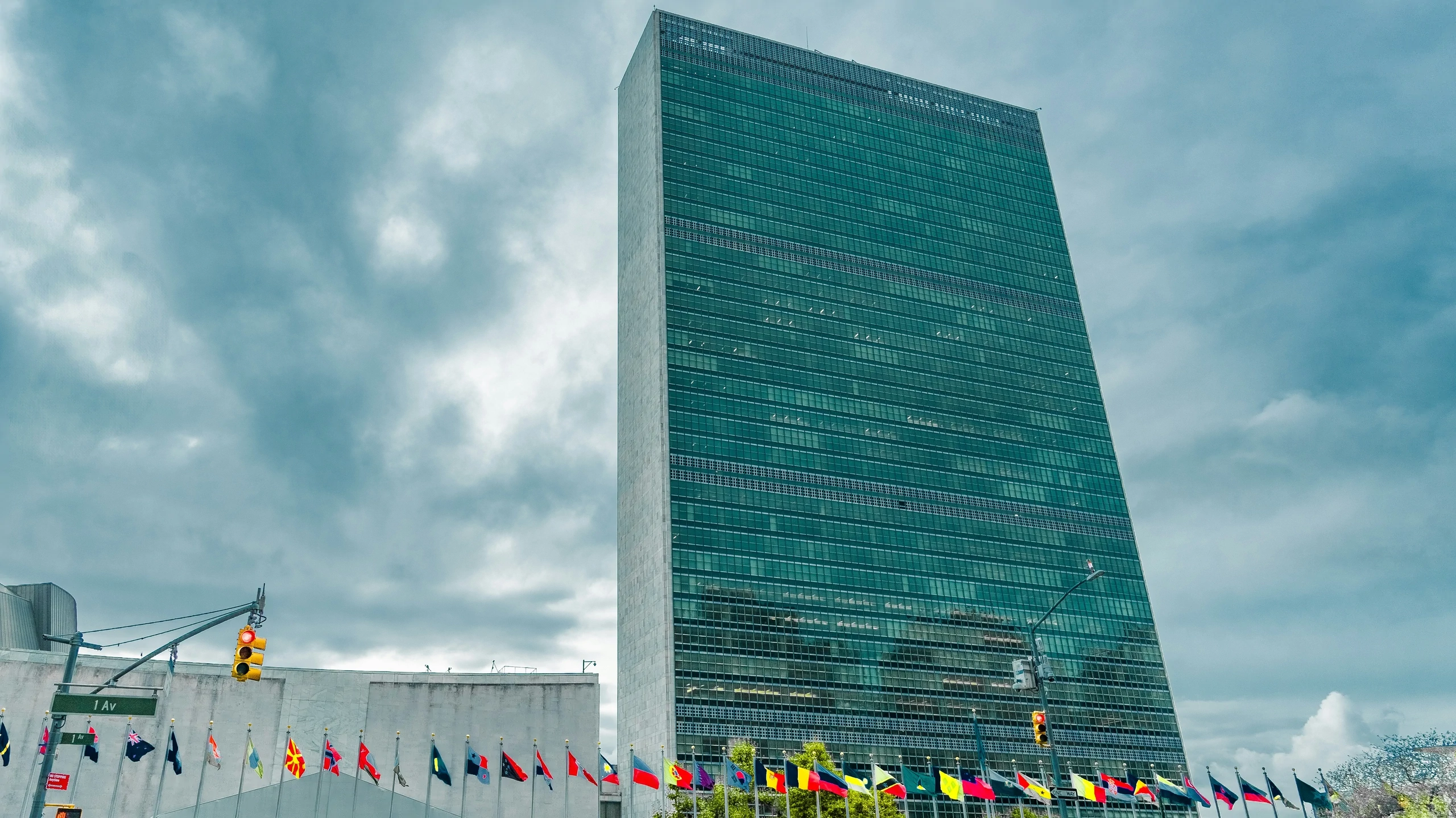 United Nations headquarters building with green-tinted glass tower and wall displaying flags of various nations.