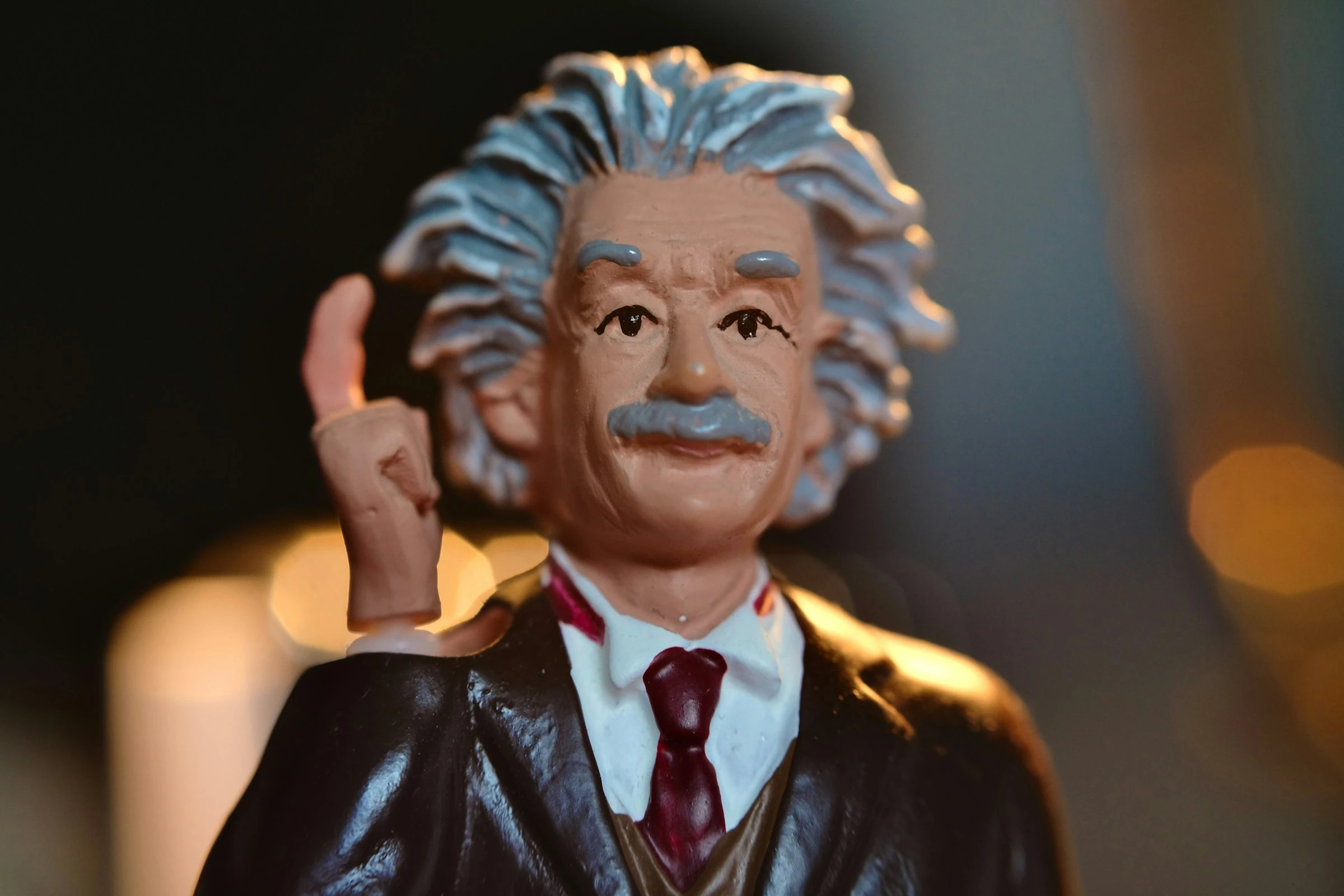 Figurine depicting Albert Einstein wearing formal black jacket, white shirt, and burgundy tie, with distinctive voluminous white hair and dark mustache, raising left hand in gesture, photographed against dark background with warm lighting.