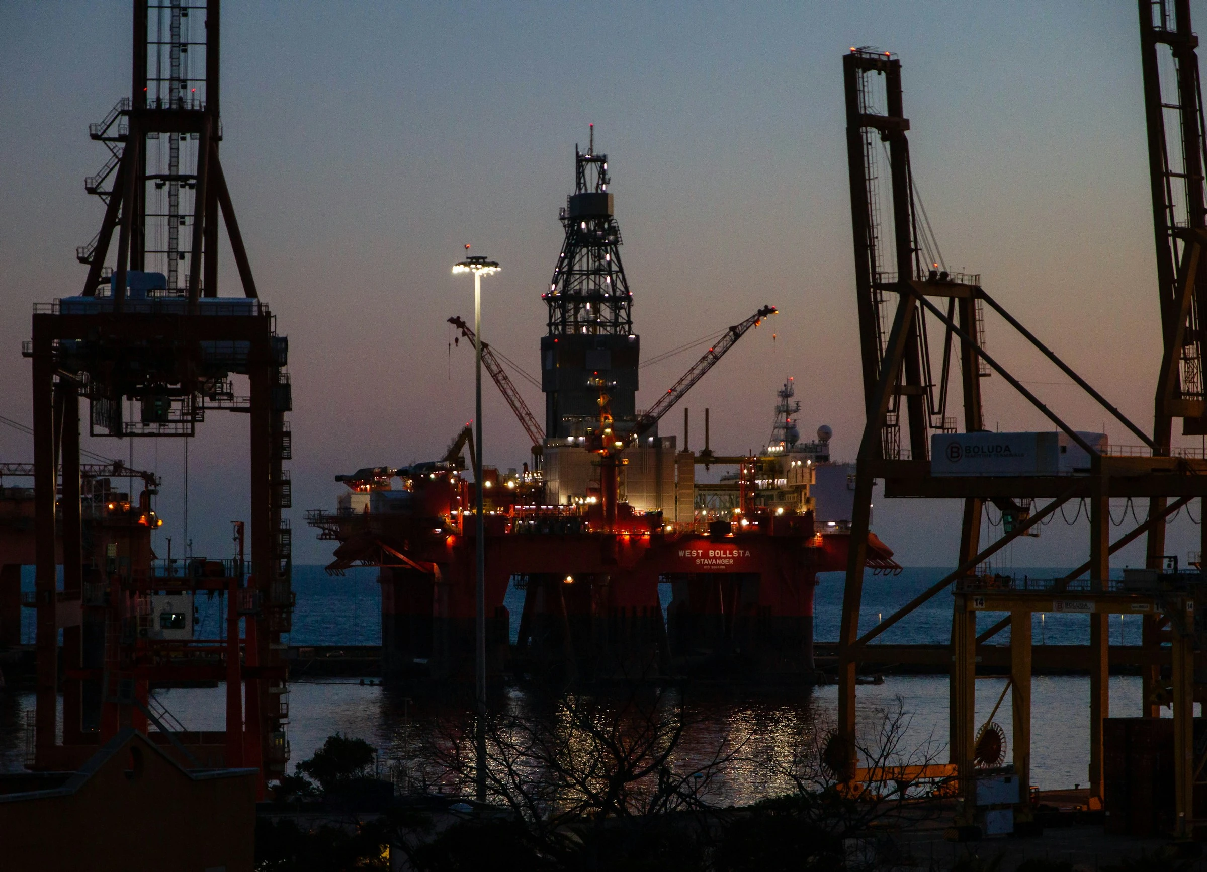 Illuminated offshore oil platform surrounded by industrial cranes at dusk, with calm water and bare tree branches in foreground.
