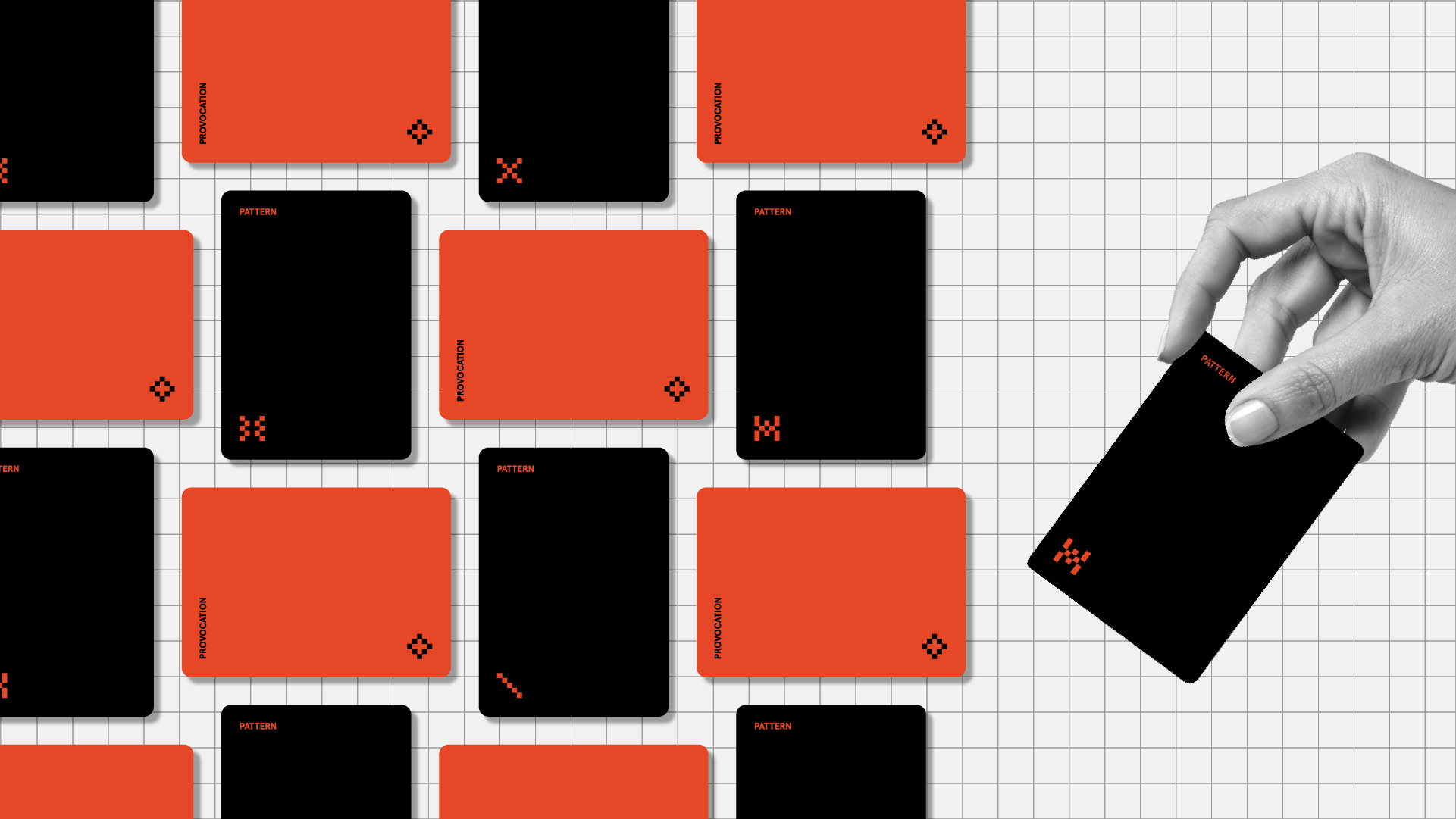 Hand picking up black card labelled "PATTERN" from grid arrangement of scattered red and black cards on white background. Red cards labelled "PROVOCATION", black cards labelled "PATTERN", each with small pixel symbols.
