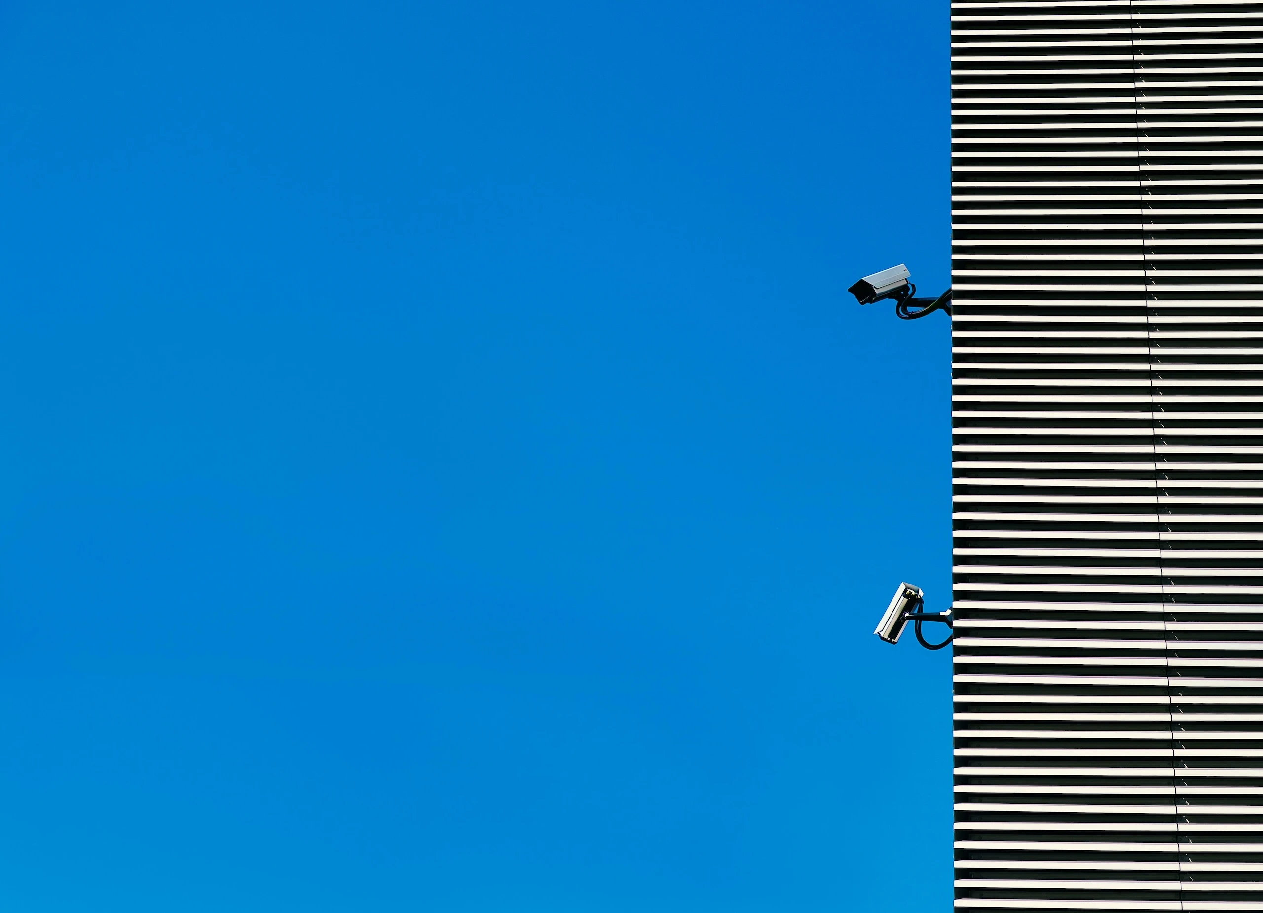 Two security cameras mounted on the corner of a horizontally slatted building, against a plain blue sky.
