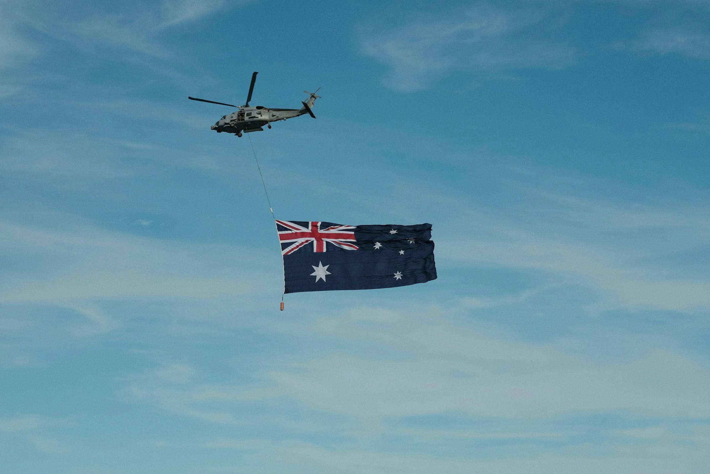 Military helicopter in flight towing a large Australian flag on a line, against a blue sky with thin cloud.