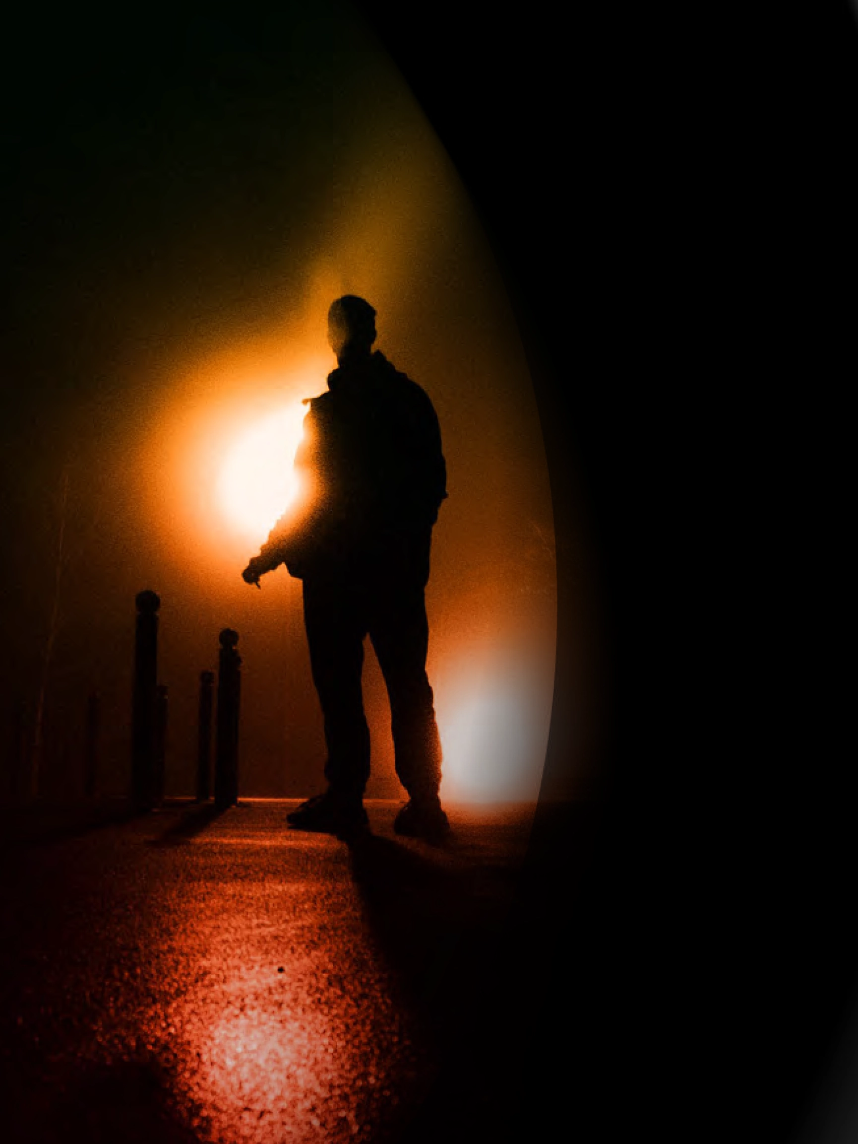 Person in silhouette standing in concrete corridor with pillars, backlit by bright orange and white glow.
