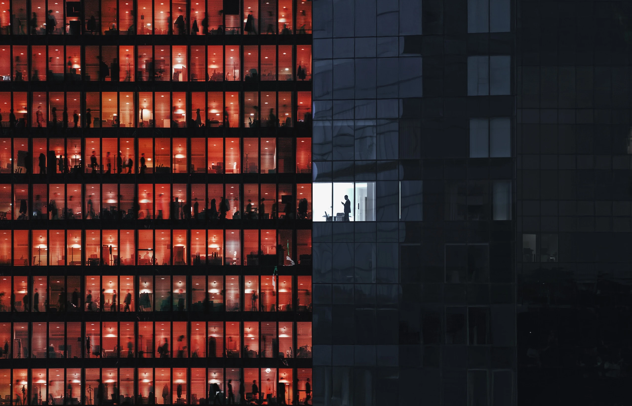Office building at night; left side has many windows lit red-orange with blurred silhouettes of people, right side is dark except one white-lit window with a person standing.