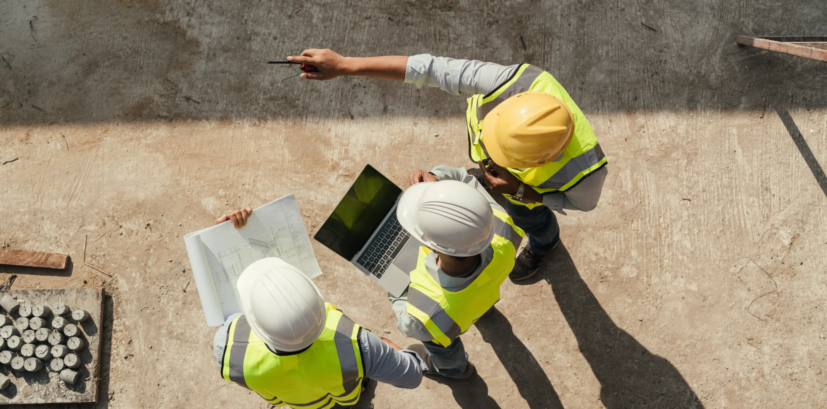 Three construction workers in yellow high-visibility vests and white hard hats standing on a concrete surface, viewed from above. One worker is pointing while the other two look at an open laptop computer and what appears to be architectural plans or blueprints spread on the ground. Construction materials including metal pipes are visible in the background.