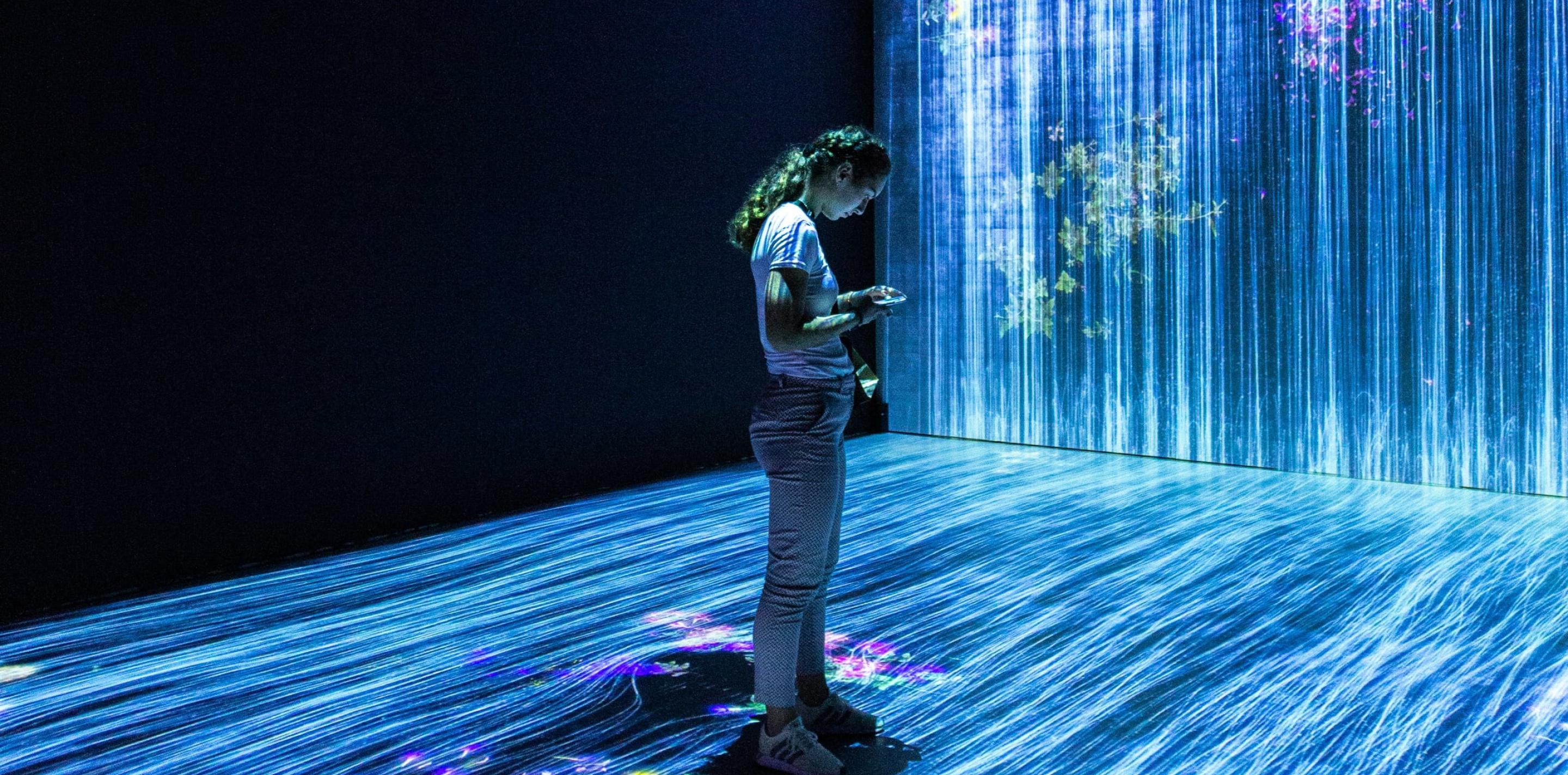 A young person interacts with their smartphone, standing on a glowing floor that projects a dynamic light display, within a dark room illuminated by a large, vivid digital art installation.