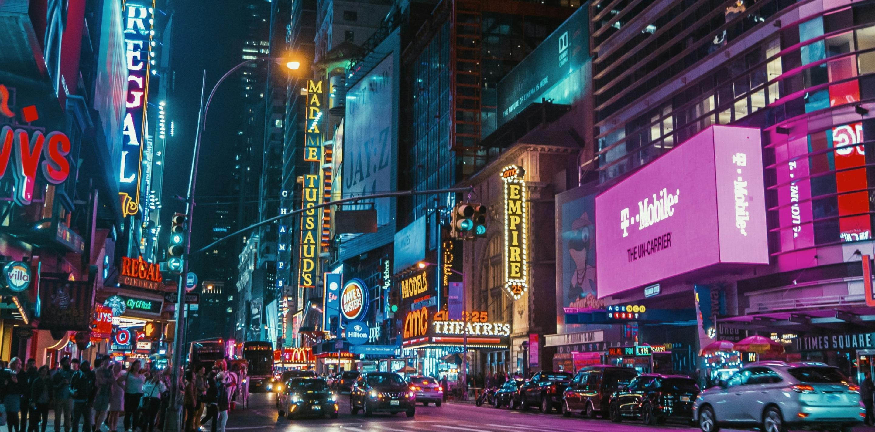 Vibrant street view of Times Square at night, bustling with traffic and pedestrians. Neon lights and billboards illuminate the scene, advertising brands like Regal Cinemas, Madame Tussauds, and T-Mobile, creating a lively urban atmosphere.