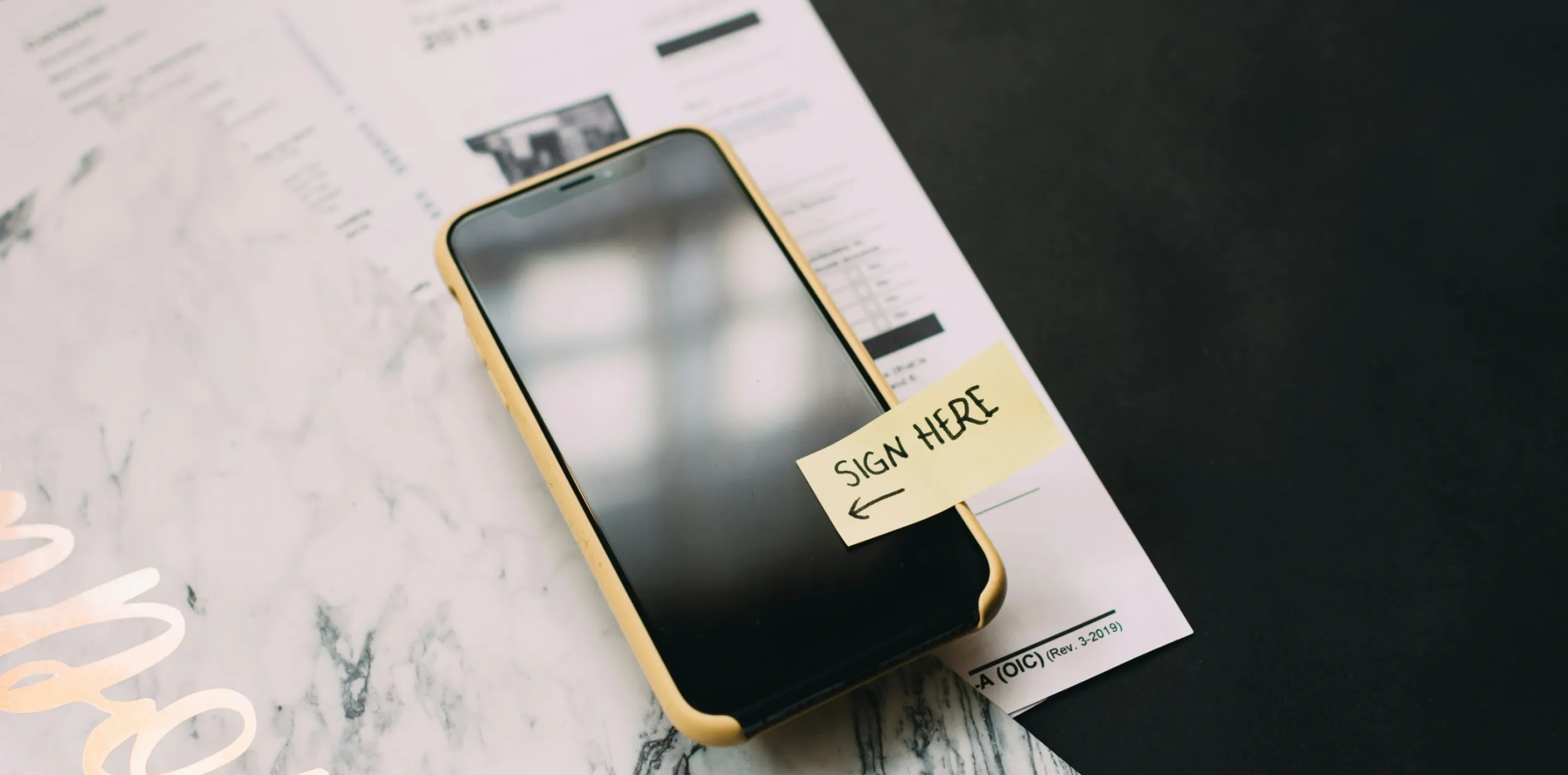 Smartphone in yellow case on printed documents with a sticky note reading "SIGN HERE" and an arrow.