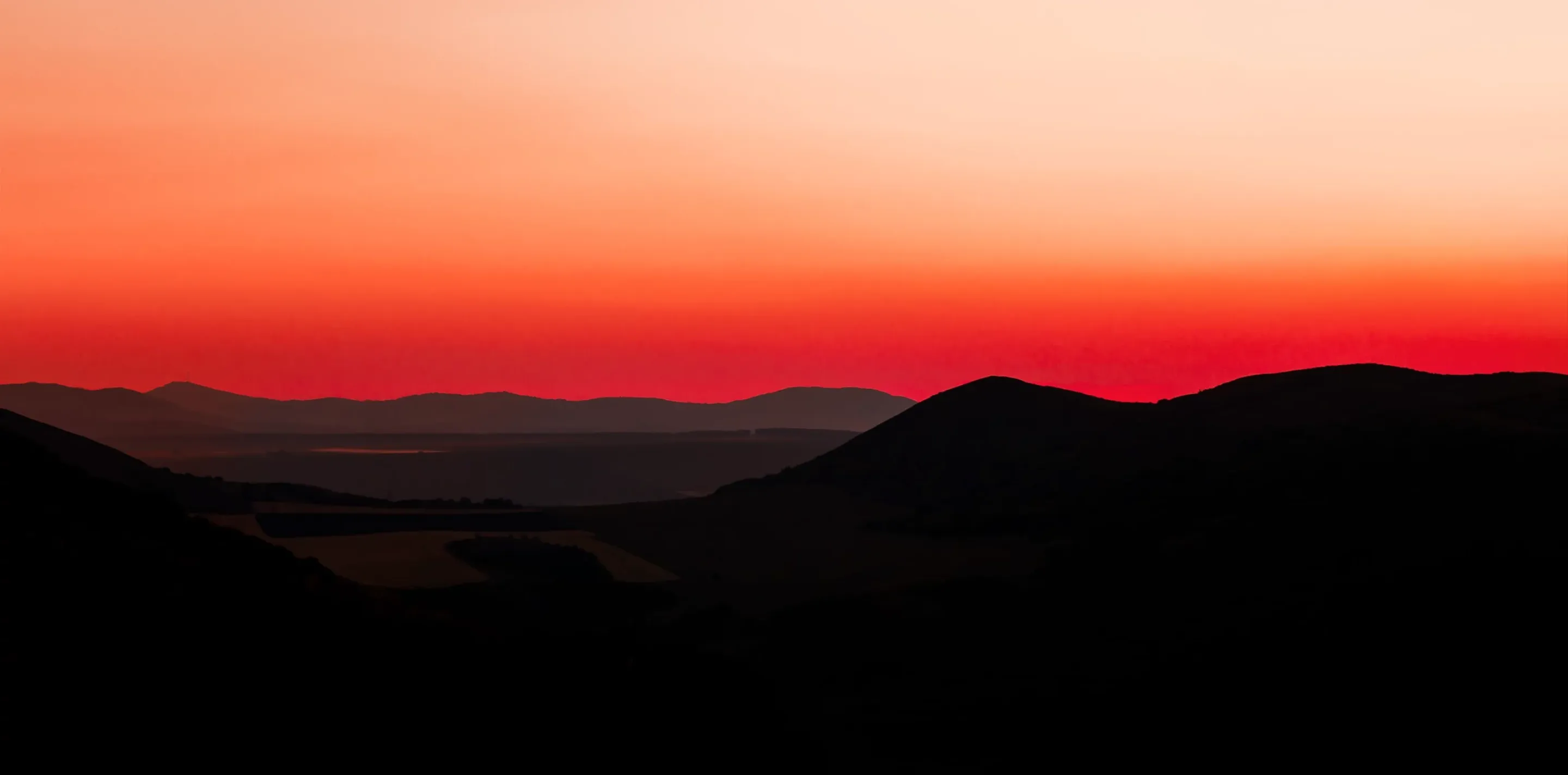 Red and orange sky at dusk with dark silhouetted hills and a valley with fields below.