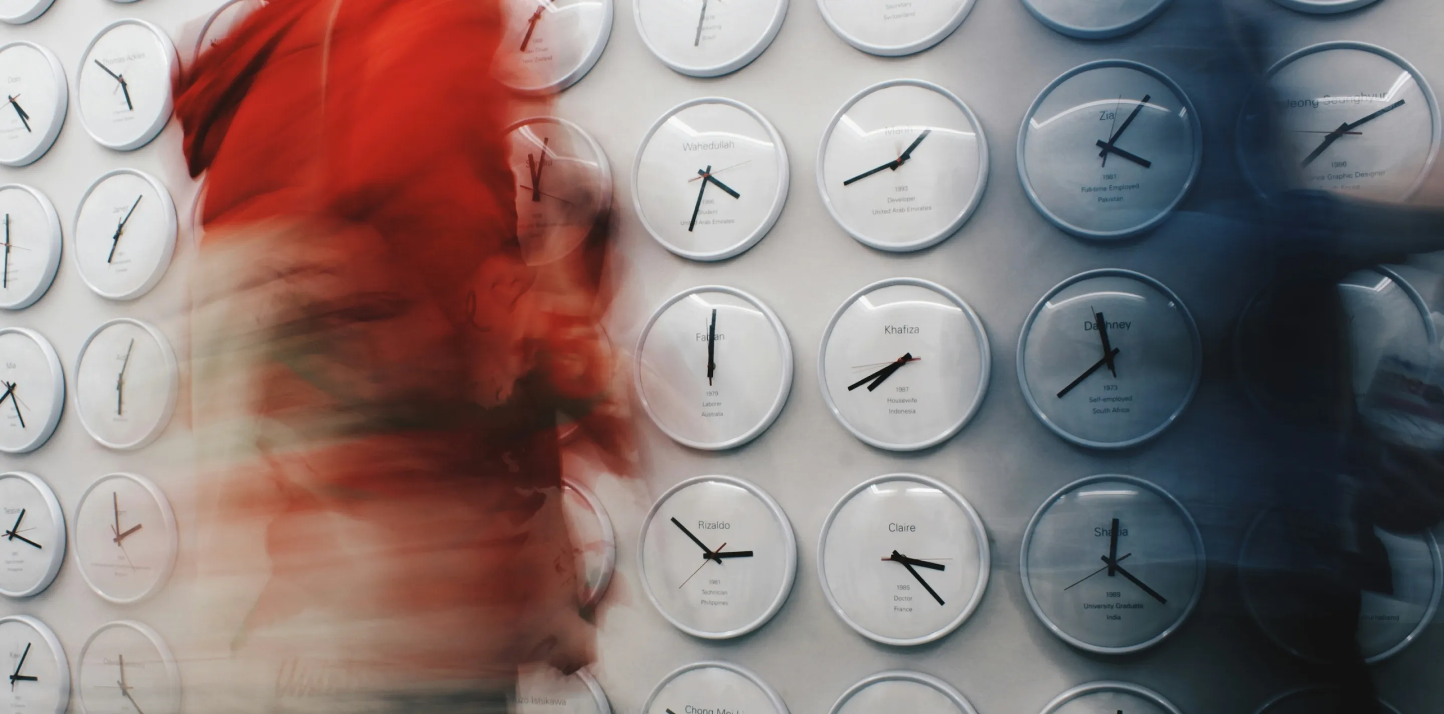 Person in red walking past a wall of white clocks, their figure blurred by motion while the clocks stay sharp.
