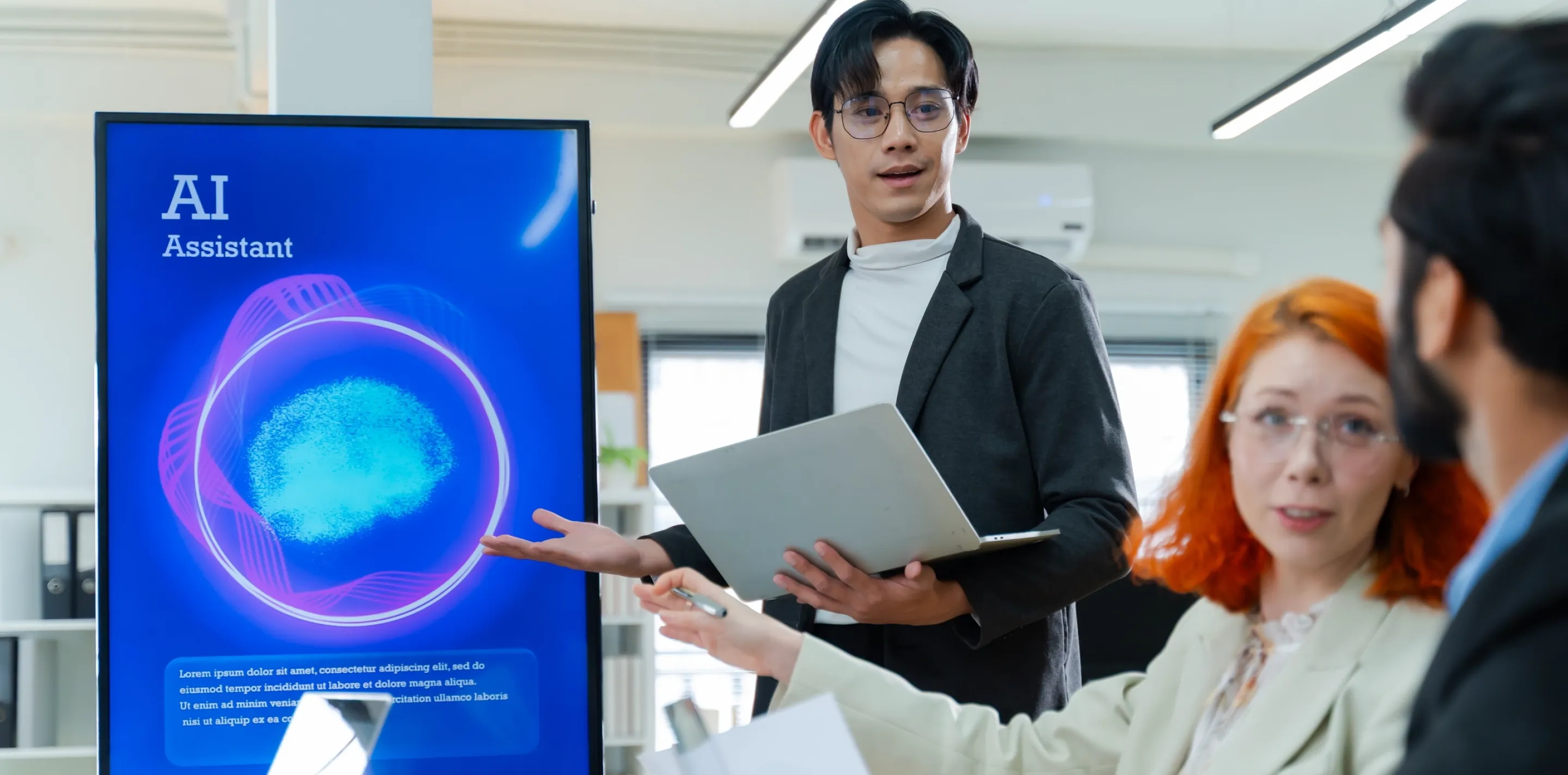 Person in blazer holding laptop presents to group in office; blue digital display showing AI Assistant logo with purple and cyan circular design on desk.