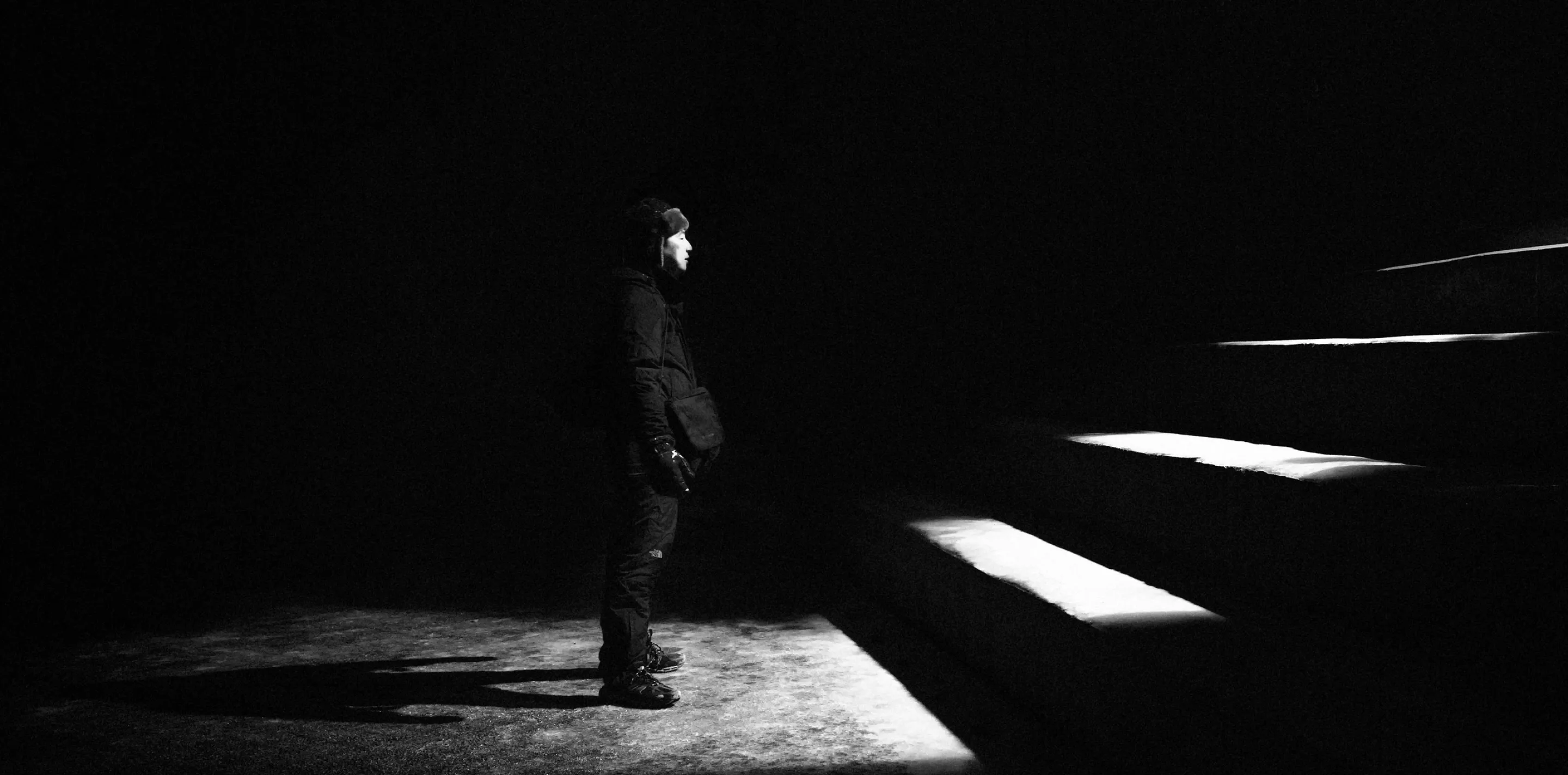 Person in winter clothing stands in darkness, face lit by a beam of light beside illuminated stair edges.
