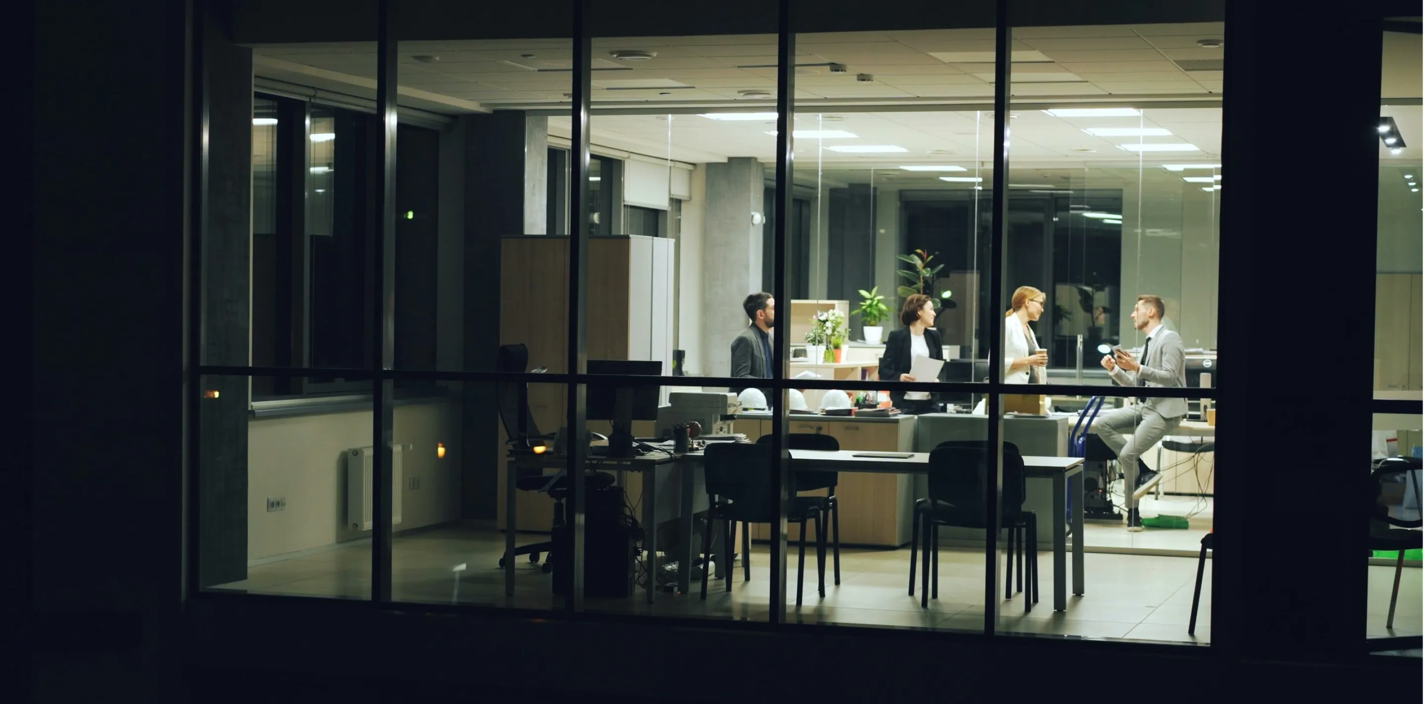 Office at night seen through windows, four people standing near desks talking.