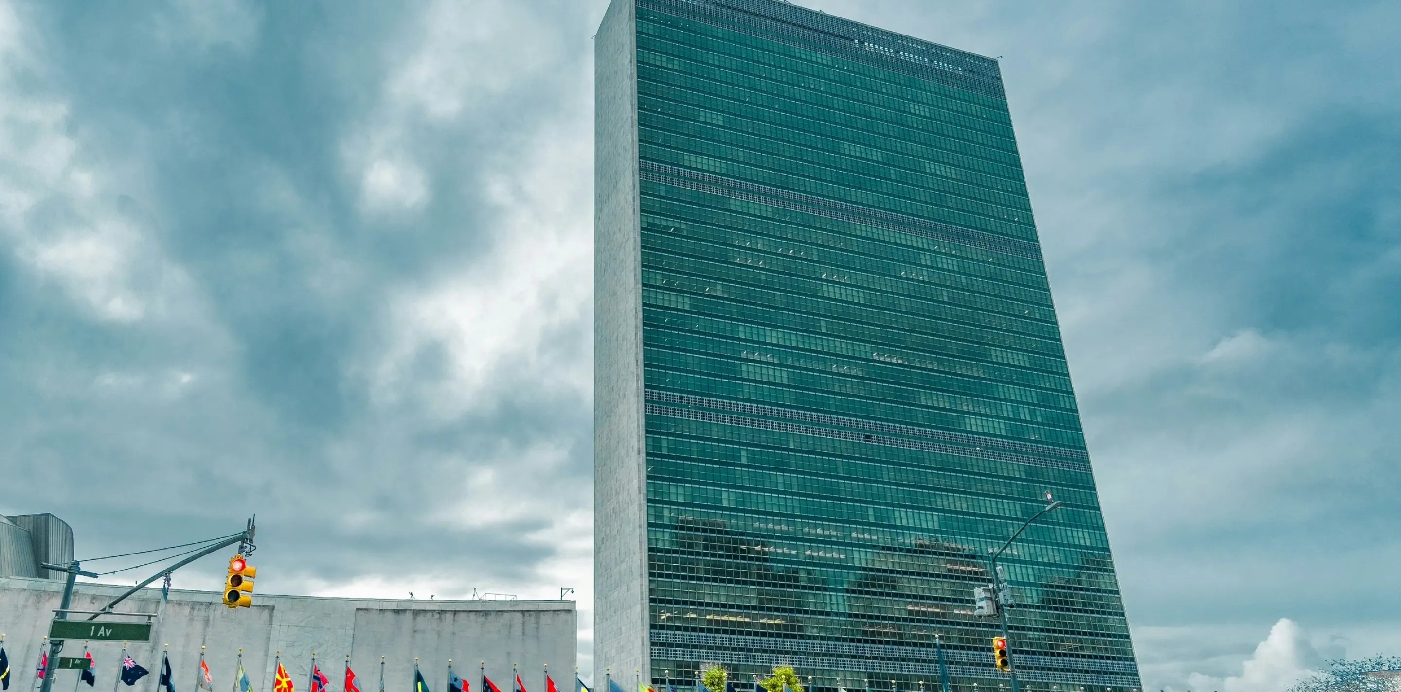 United Nations headquarters building with green-tinted glass tower and wall displaying flags of various nations.