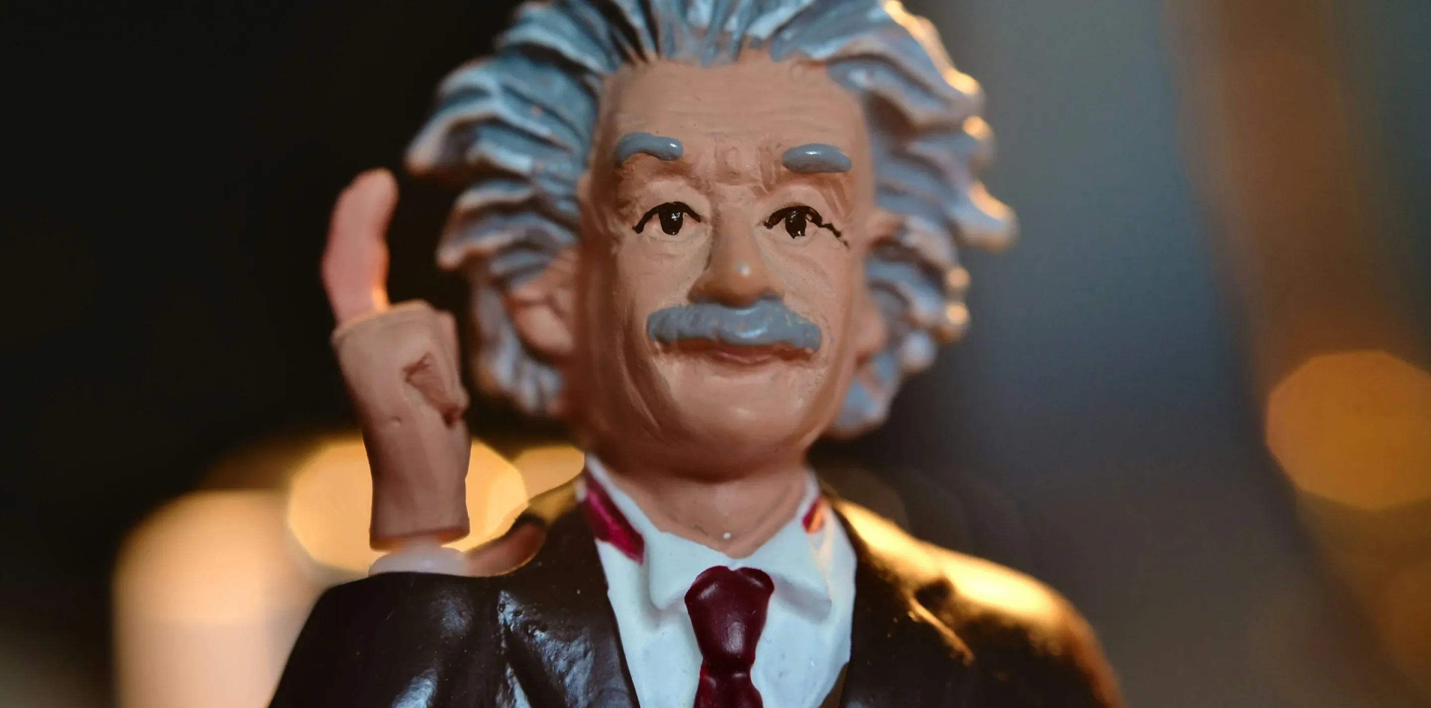 Figurine depicting Albert Einstein wearing formal black jacket, white shirt, and burgundy tie, with distinctive voluminous white hair and dark mustache, raising left hand in gesture, photographed against dark background with warm lighting.
