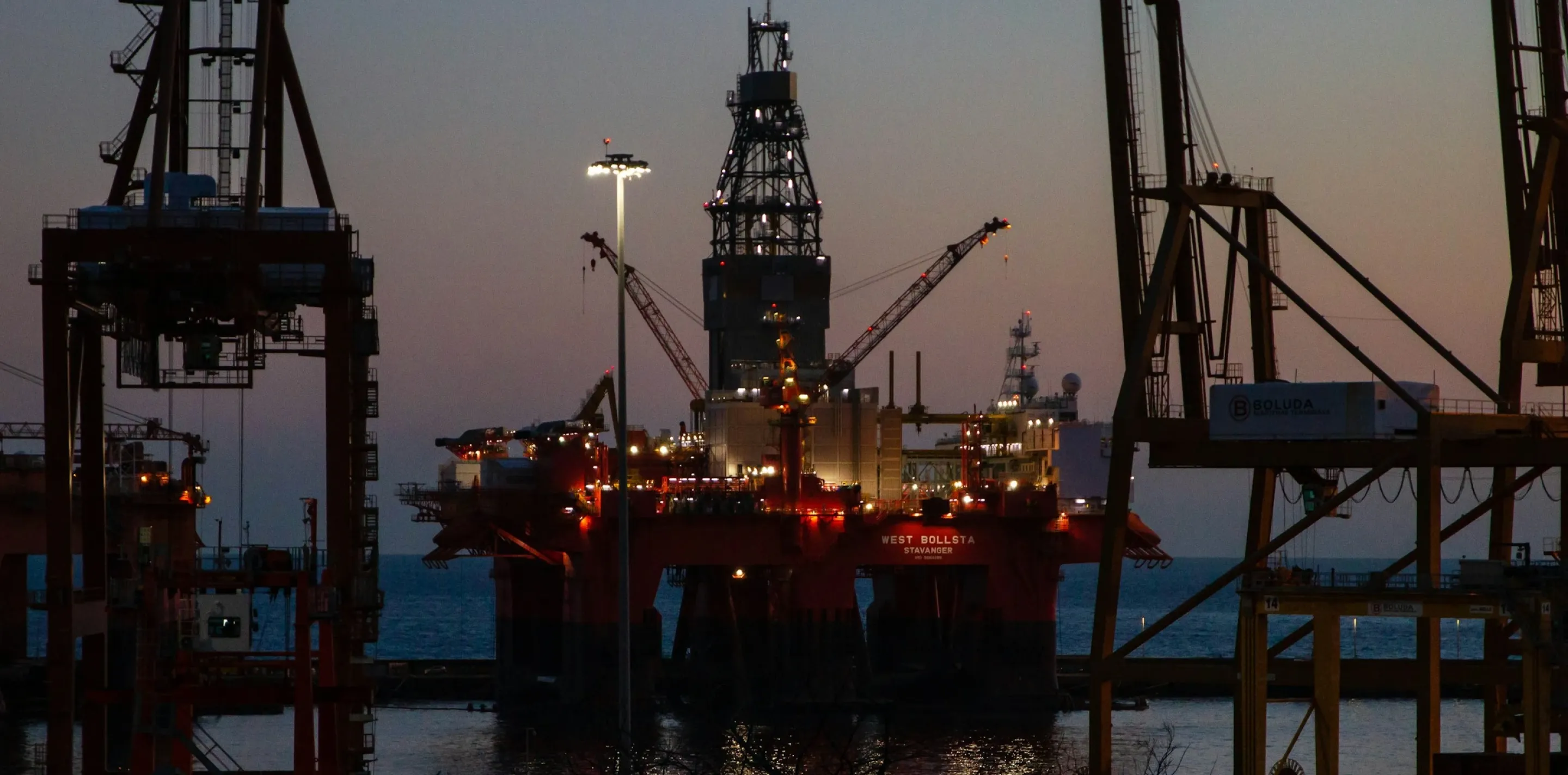 Illuminated offshore oil platform surrounded by industrial cranes at dusk, with calm water and bare tree branches in foreground.