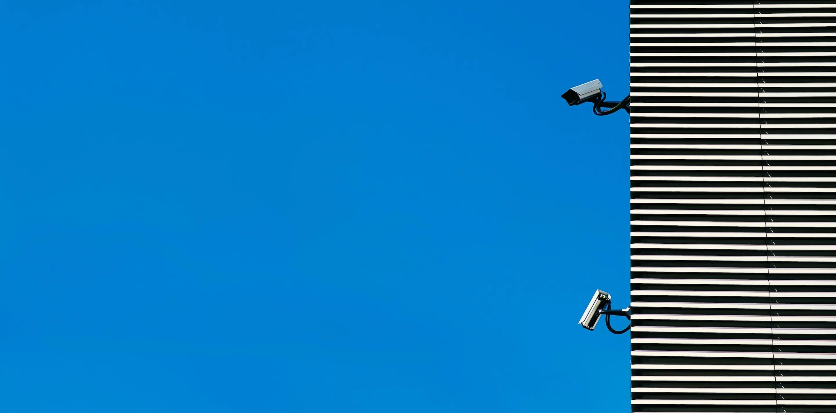Two security cameras mounted on the corner of a horizontally slatted building, against a plain blue sky.