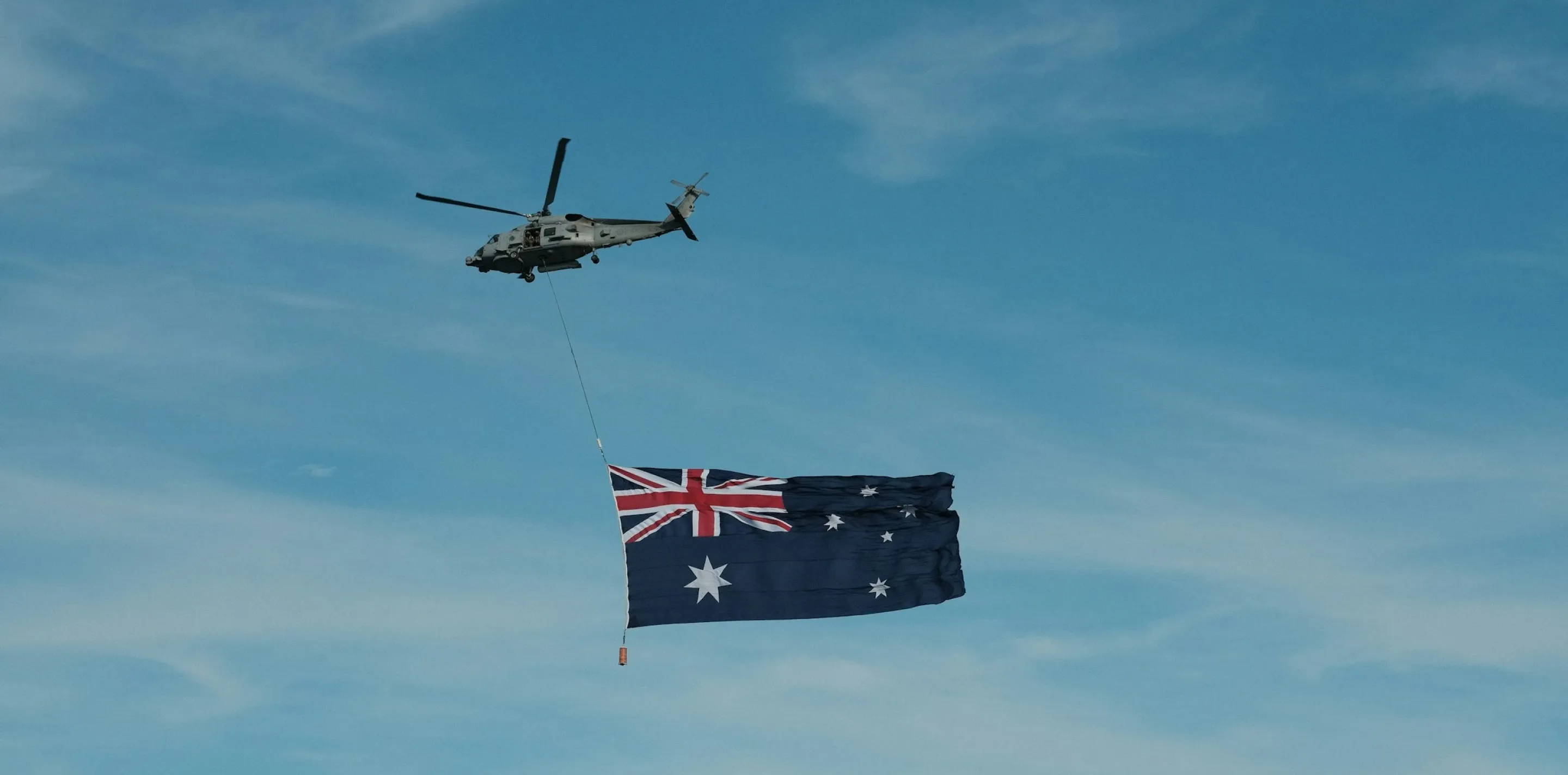 Military helicopter in flight towing a large Australian flag on a line, against a blue sky with thin cloud.