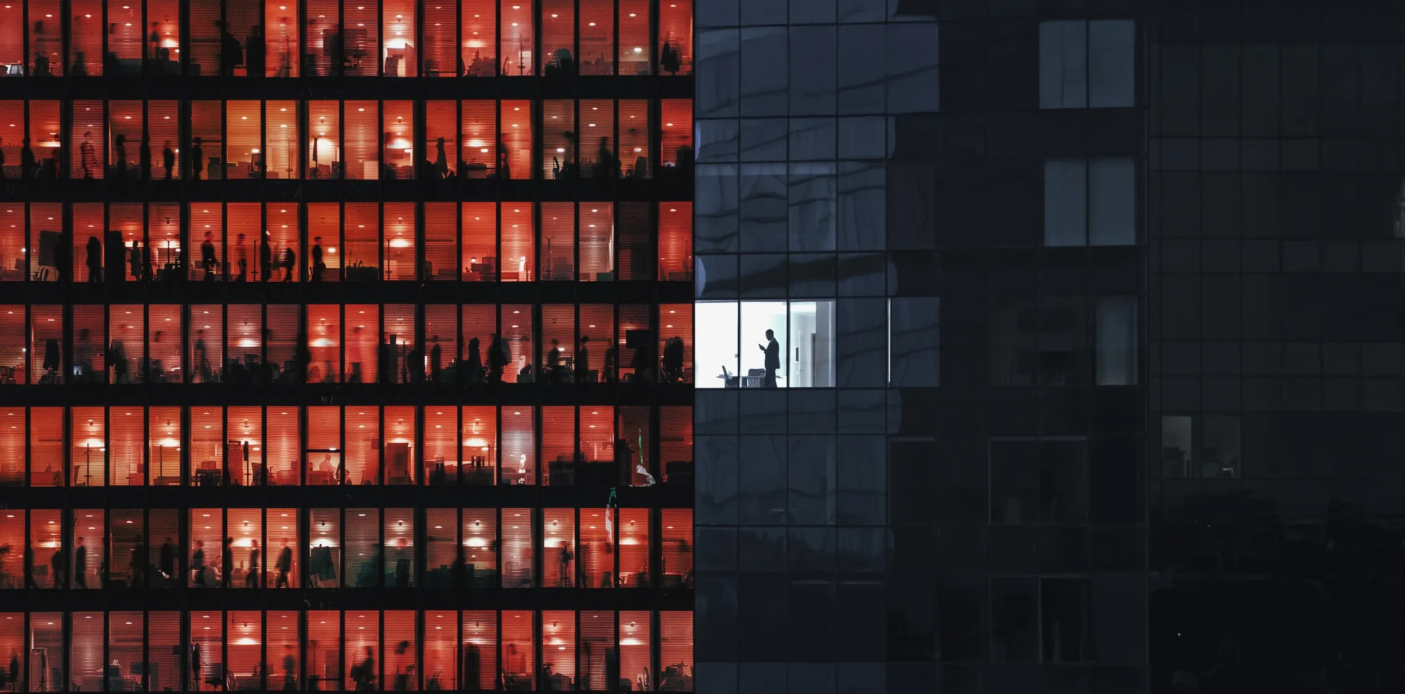 Office building at night; left side has many windows lit red-orange with blurred silhouettes of people, right side is dark except one white-lit window with a person standing.