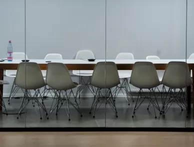 Modern conference room with a long wooden table and a combination of white and taupe Eames-style chairs reflected in a glass partition, a solitary water bottle and notepad on the table indicating a recent or upcoming meeting.