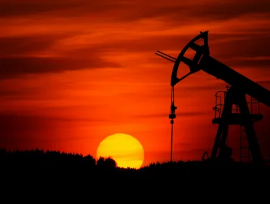 Oil pump jack silhouetted against a red and orange sunset sky, with the sun setting behind a dark treeline.