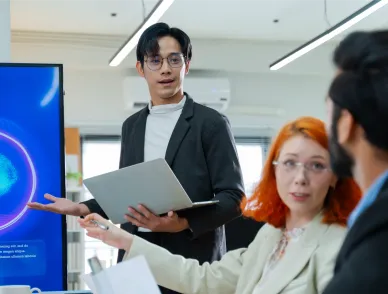 Person in blazer holding laptop presents to group in office; blue digital display showing AI Assistant logo with purple and cyan circular design on desk.