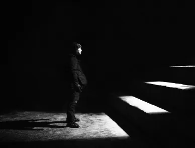 Person in winter clothing stands in darkness, face lit by a beam of light beside illuminated stair edges.