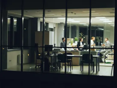 Office at night seen through windows, four people standing near desks talking.