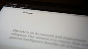 A close-up view of a tablet screen displaying the OpenAI website with the text, 'OpenAI is an AI research and deployment company. Our mission is to ensure that artificial general intelligence benefits all of humanity.' The screen is partially blurred, focusing on the OpenAI logo and introductory text.