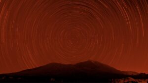 A long-exposure photograph showing star trails in circular patterns above a dark mountain range, with a red-orange hue tinting the entire scene.