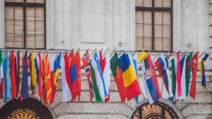 A row of international flags from various countries is displayed in front of a stone building with large windows.