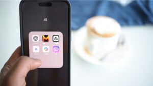 A person holds a smartphone displaying a folder labeled “AI” with six AI-related apps: ChatGPT, Mistral AI, Claude, Gemini, Copilot, and Poe, in front of a blurred background featuring a cappuccino on a table.