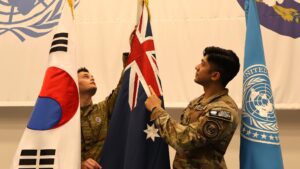 Two uniformed military personnel raise the Australian flag between the Republic of Korea and United Nations flags.